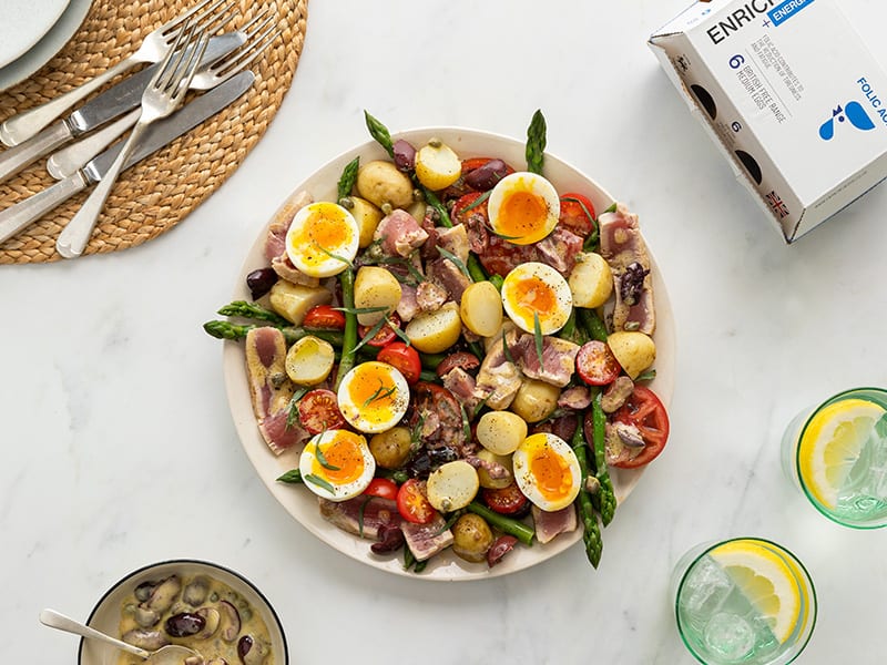 Tuna Niçoise salad on a large platter surrounded by glasses of water, cutlery and a white box of Enriched Eggs