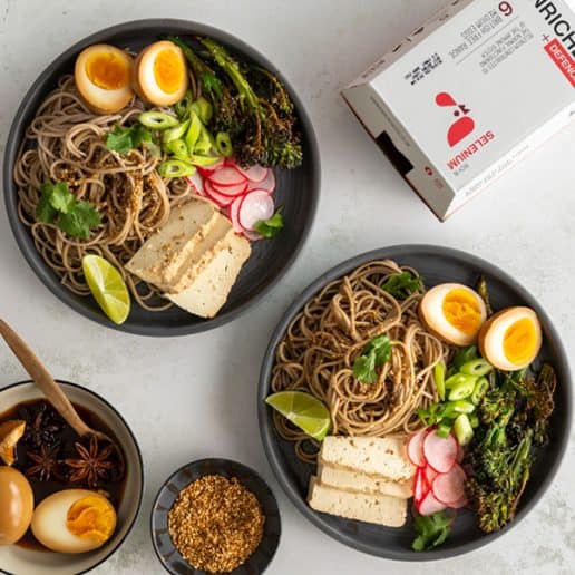 Two plates of soba noodles with sliced boiled eggs, tofu and vegetables, next to a box of eggs.