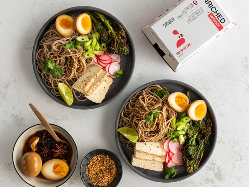 Two plates of soba noodles with sliced boiled eggs, tofu and vegetables, next to a box of eggs.