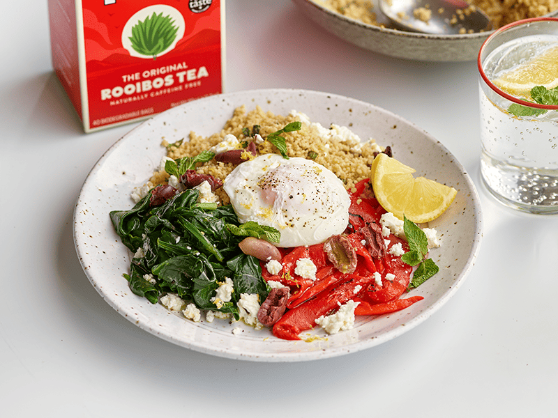 Poached egg sits on top of a bowl of chopped vegetables and lemon wedge, in front of a red box of Tick Tock Original Rooibos tea and glass of water