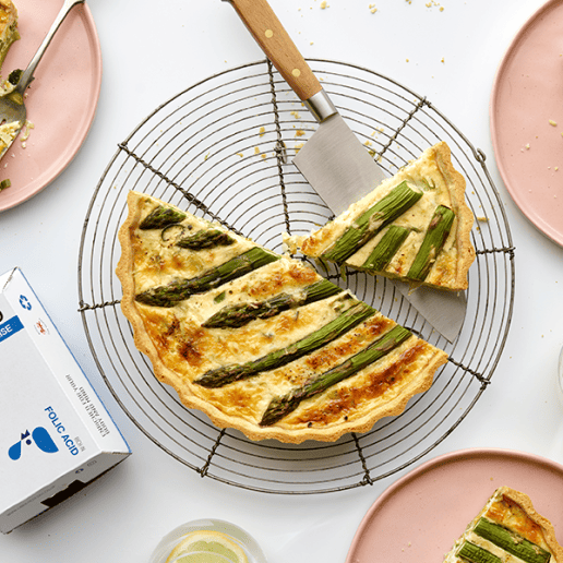 Quiche topped with asparagus in a wire cooling rack, surrounded by plates with slices of quiche and a box of Enriched eggs.