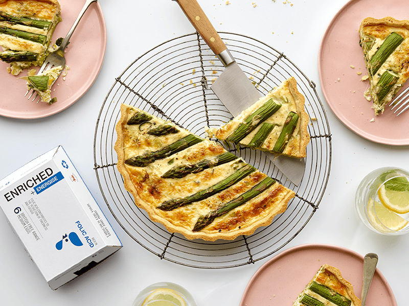 Quiche topped with asparagus in a wire cooling rack, surrounded by plates with slices of quiche and a box of Enriched eggs.