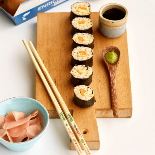 Six sushi rolls on a wooden board, next to wooden chopsticks, a small bowl of soy sauce, a dollop of wasabi and small bowl of pickled ginger.