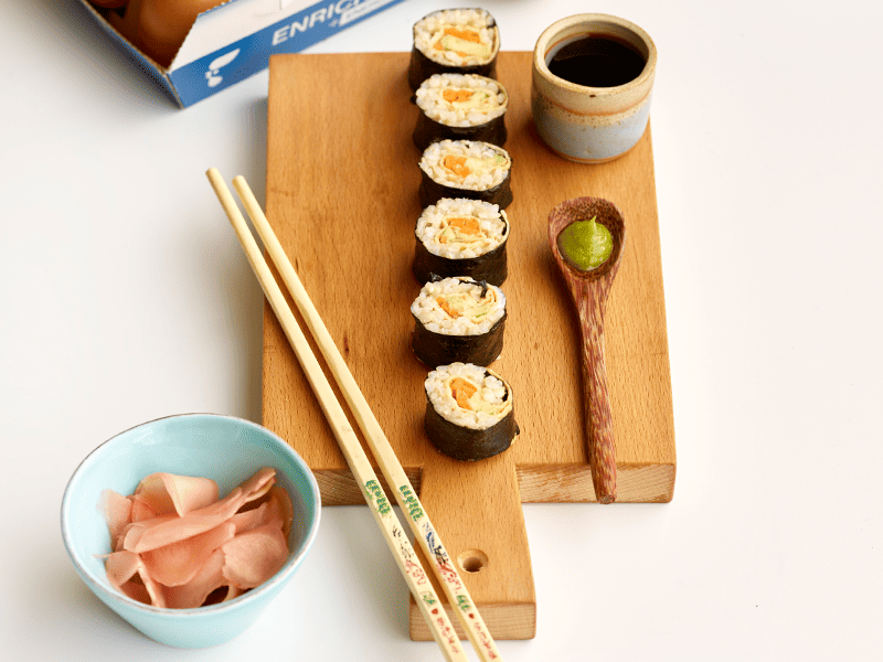 Six sushi rolls on a wooden board, next to wooden chopsticks, a small bowl of soy sauce, a dollop of wasabi and small bowl of pickled ginger.