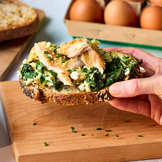 Soft baked eggs with spinach and herbs sit on a slice of crunchy wholemeal toast, topped with shreds of hot salmon. The toast is being lifted off a wooden board by a woman's hand. In the background of the photo there are eggs in a carton and the rest of the load of bread.