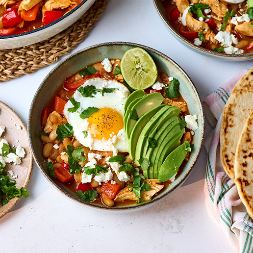 A bowl full of chicken, peppers, onions and cannellini beans in a tomato sauce topped with a fried egg, sliced avocado and a lime wedge sits on a white table. The dish has a scattering of coriander across the top.