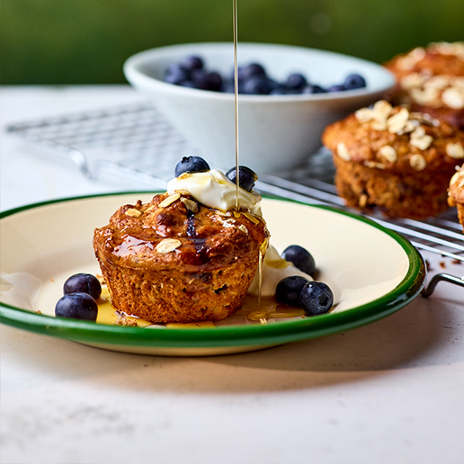 A whole peanut butter, oat and blueberry muffin sits on a white and green plate surrounded by six whole blueberries. The muffin has a stream of maple syrup being drizzled on it from above, some of it pooling in the base of the plate. The muffin is also topped with a dollop of greek yogurt. There are more muffins and a bowl of blueberries sitting on a wire cooling rack in the background of the photo.