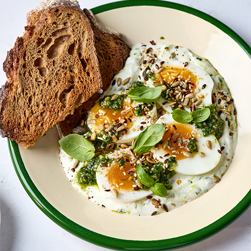 In a white bowl rimmed in green, there is yogurt mixed with pesto and topped with four halves of boiled eggs, the yolk facing up. Sprinkled across the top of the whole dish are pine nuts and basil leaves. On side of the bowl is two slices of toasted wholemeal bread.