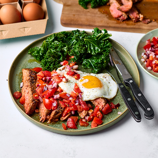 A round green plate sits on a white table. On the plate is sliced pieces of steak, tomato salsa, a fried egg and kale. Extra eggs, steak and salsa surround the plate on the table.