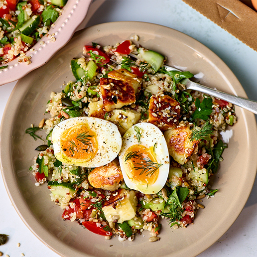 A light brown bowl filled with a mixture of quinoa, seed-crusted halloumi, chopped tomatoes, cucumber, spring onions and herbs, topped with a halved jammy egg.