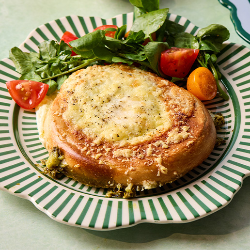 This air fryer cheesy pesto egg bagels is sitting on a round, stripy green and white plate with some salad greens and tomatoes sitting on the side.