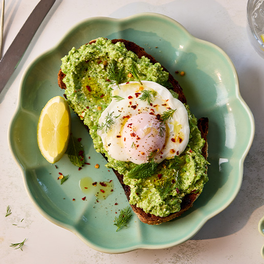 Crushed Pea toasts with broad beans, cottage cheese and poached eggs on top, served on a green leaf shaped plate with a wedge of lemon to the left hand side and sprinkled with chilli flakes.