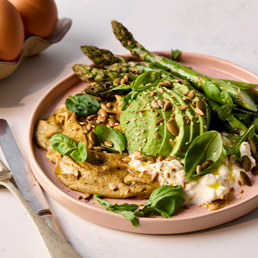 The green breakfast plate includes folded pesto eggs, grilled asparagus, avocado and cottage cheese, all served on a pale pink plate with a knife sitting to the left hand side